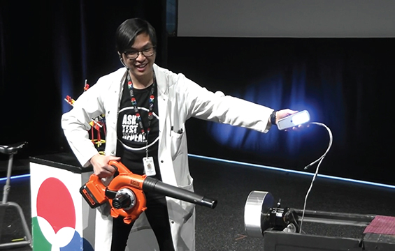 Un éducateur en sciences portant un sarrau fait une démonstration à l'aide d'un souffleuse à feuilles et d'une lampe allumée.
