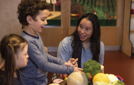 Une femme et deux enfants jouent avec de faux aliments dans un décor de supermarché.